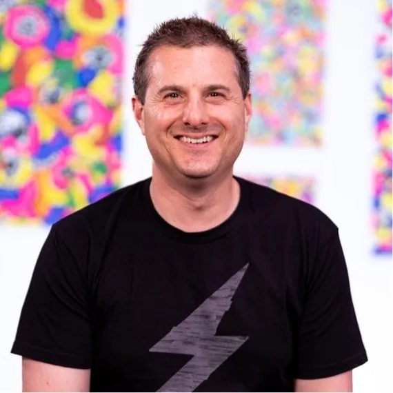 Avi Spivack smiling in a black t-shirt with a lightning bolt, in front of Nataly's colorful paintings