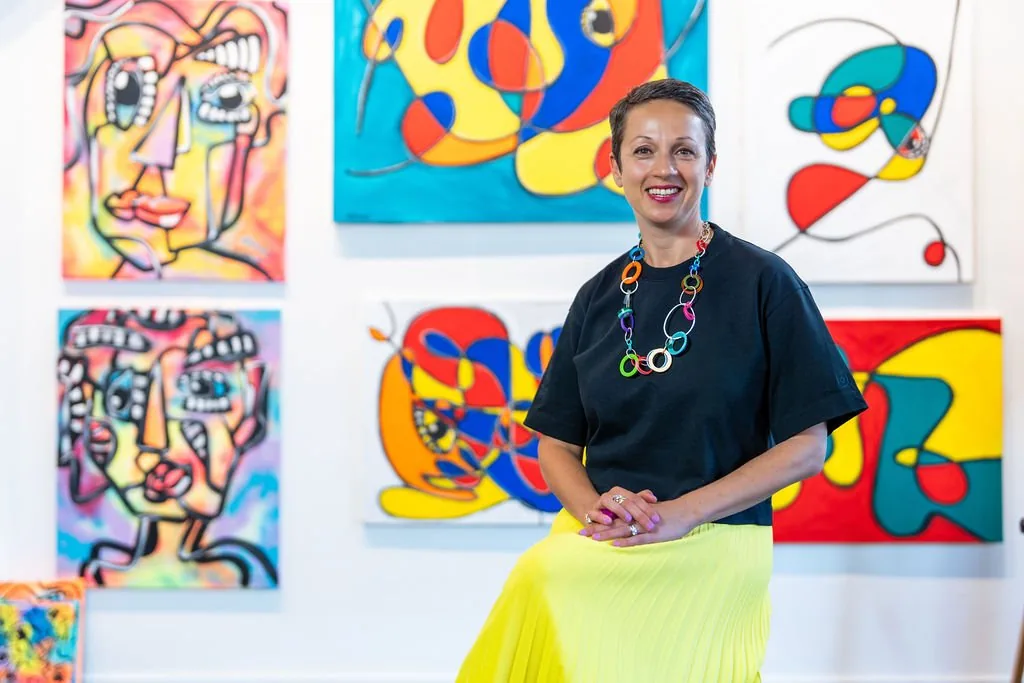 Nataly Kogan smiling in a yellow skirt, seated in front of a wall of her colorful paintings